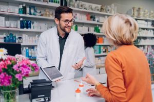 Young male caucasian pharmacist showing tablet to senior female customer in a pharmacy with female pharmacist in the background. Shelves with health care products.